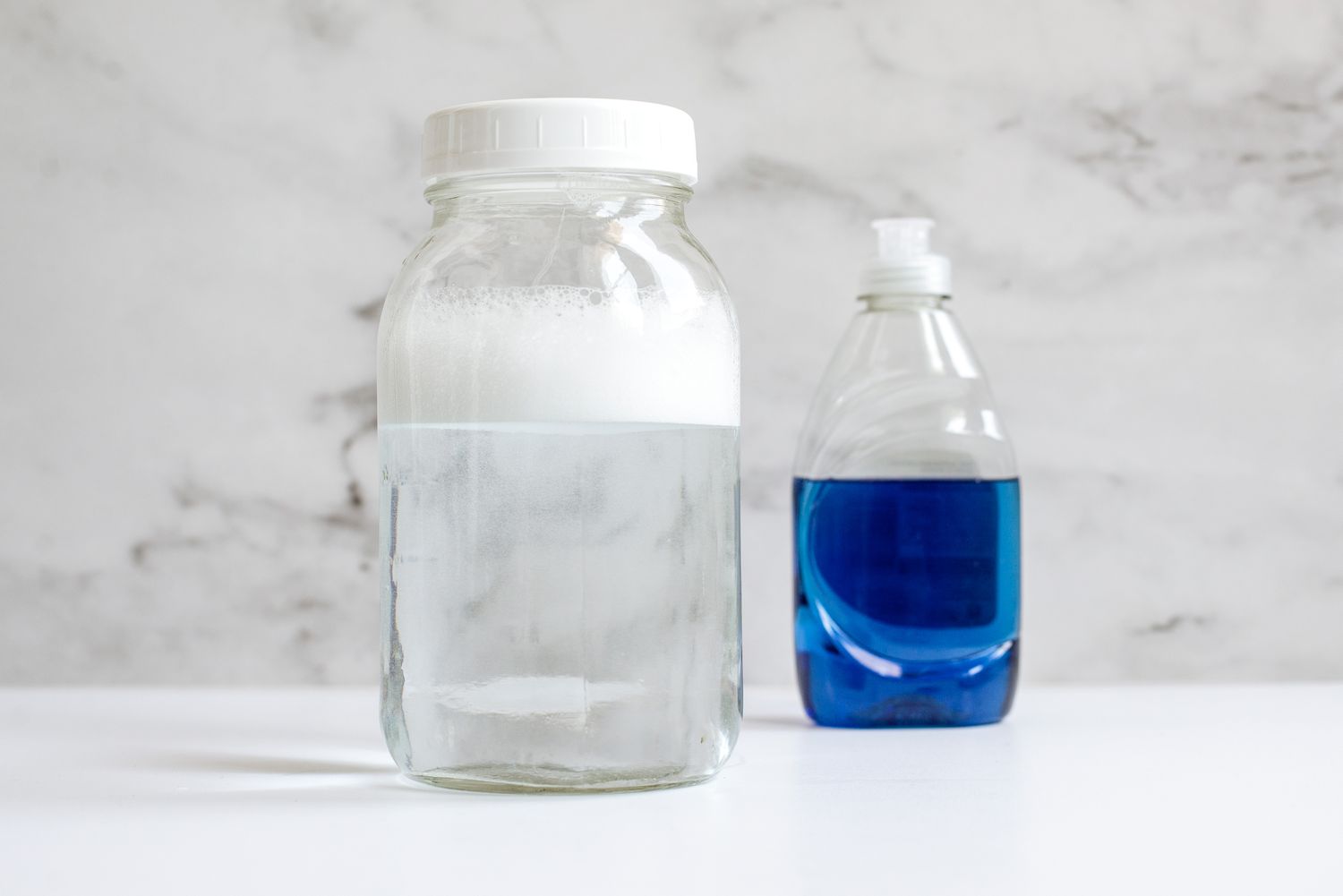 A sizable transparent container filled with water and soap bubbles positioned in front of a bottle of liquid dish soap.