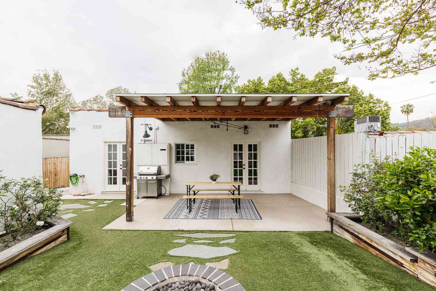 A wooden pergola connected to a white residence provides shade over a table and grill in the backyard.